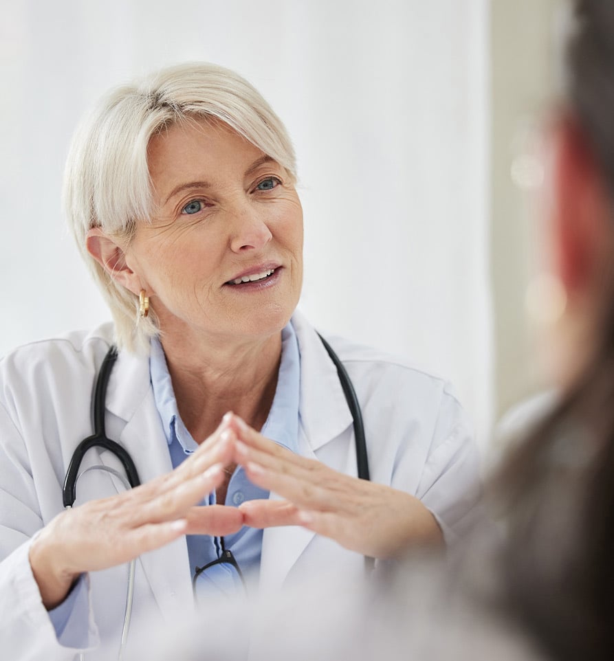 Eine Ärztin in weißem Kittel und mit Stethoskop, die während eines Gesprächs mit einem Patienten aufmerksam zuhört und die Hände vor der Brust gefaltet hat.