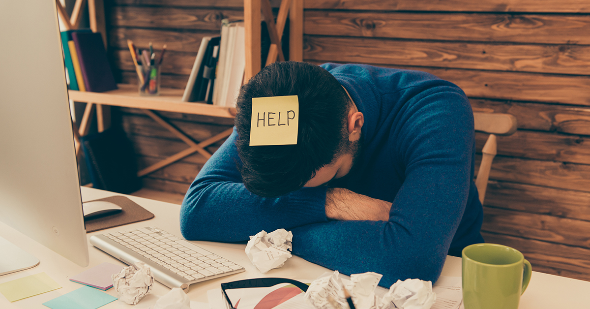 Psychische Gesundheit am Arbeitsplatz: Ein Mitarbeiter sitzt mit verschränkten Armen und gesenktem Kopf an seinem Schreibtisch und hat einen Pos-it mit der Aufschrift „Help“ auf dem Kopf kleben