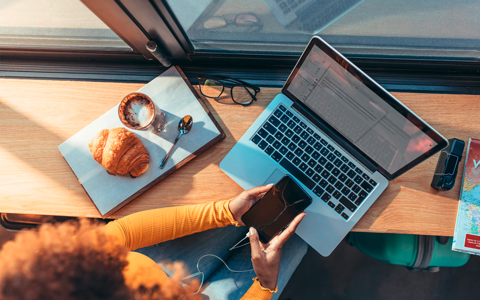 Eine Frau, die auf ihrem Smartphone rumtippt, dabei den Laptop aufgeklappt hat und ein Croissant und Kaffe neben sich stehen hat