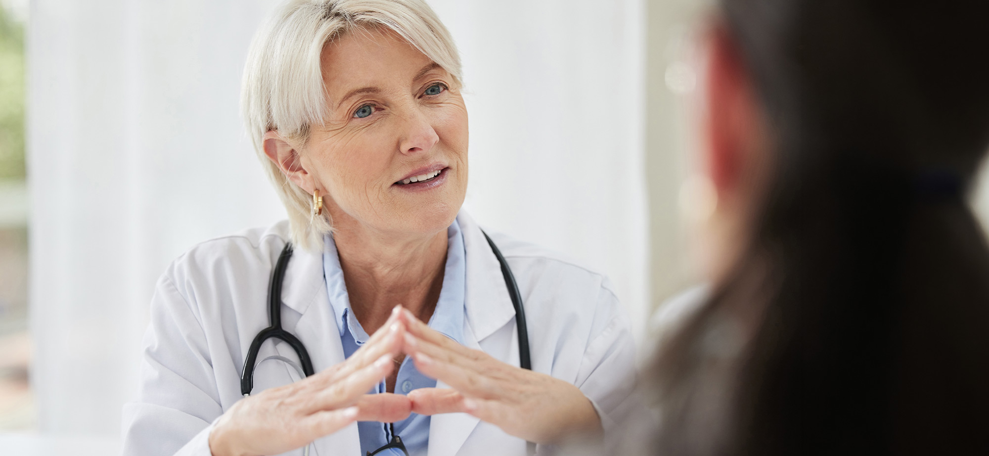 Eine Ärztin in weißem Kittel und mit Stethoskop, die während eines Gesprächs mit einem Patienten aufmerksam zuhört und die Hände vor der Brust gefaltet hat.